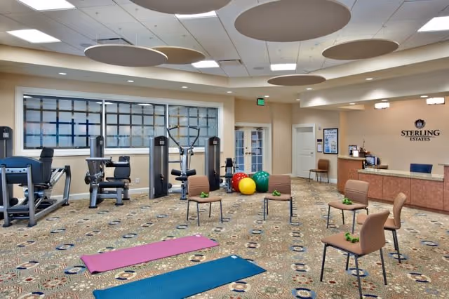 Bright interior exercise room with cardio and strength machines, yoga mats, chairs, and colorful exercise balls near a reception desk.