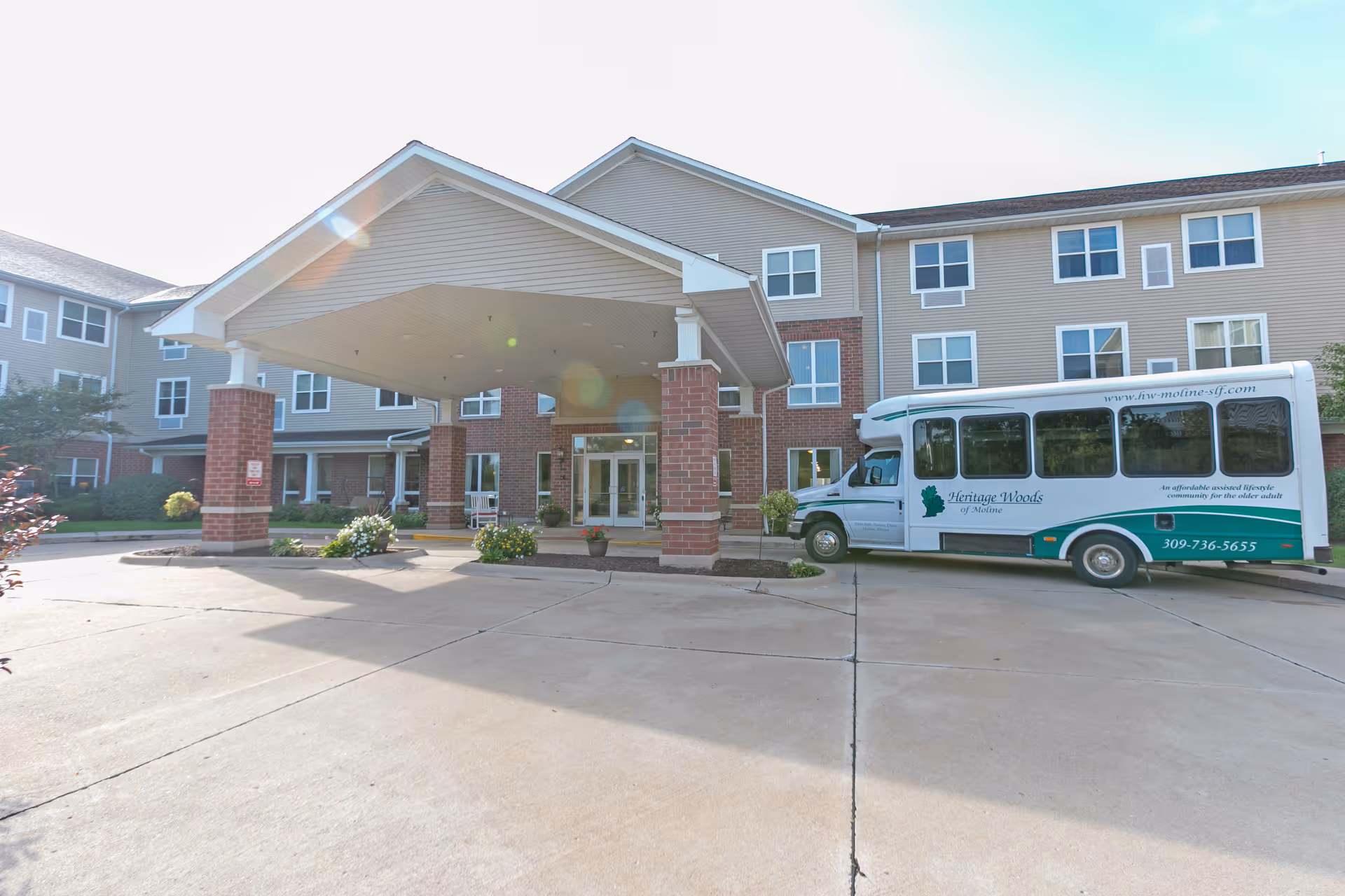 Front exterior view of a senior living facility named Heritage Woods of Moline, featuring a covered entrance supported by brick columns and a shuttle bus parked to the right with the facility's name and contact information.