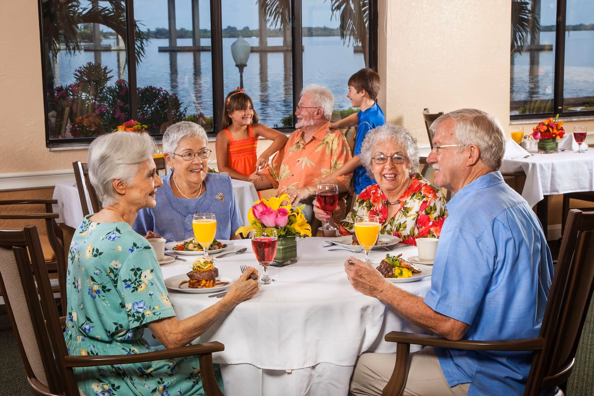 A group of elderly people and two children enjoying a meal together at a dining table in a bright room with large windows overlooking a waterfront view. The table is set with plates of food, glasses of orange juice and red wine, and a floral centerpiece.