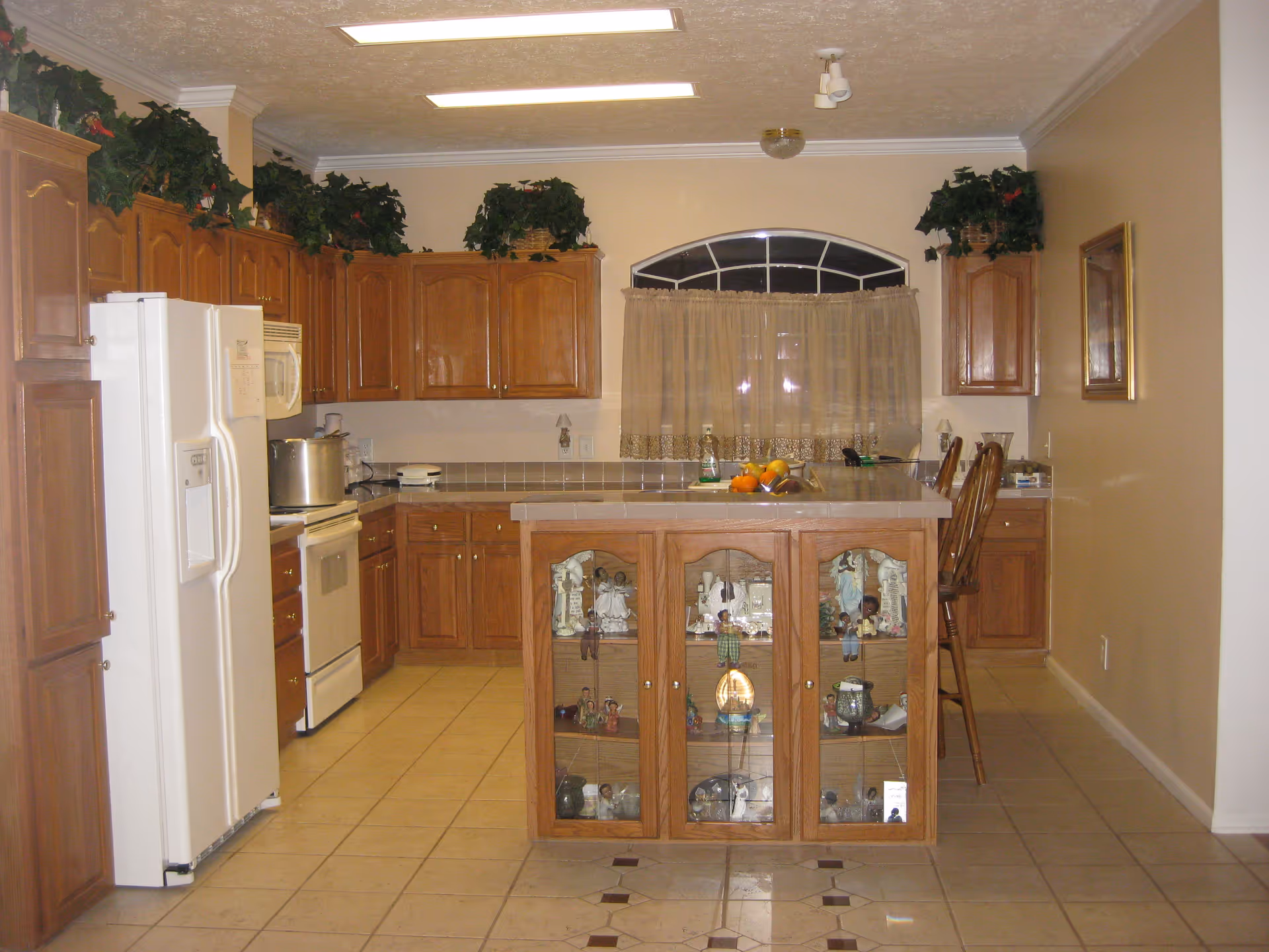 A kitchen with wooden cabinets and a white refrigerator on the left. There is a kitchen island with glass-front cabinets displaying decorative items. The countertop has a bowl of fruit and some other items. Above the cabinets are green plants. The floor is tiled, and there is a window with a curtain in the back wall.