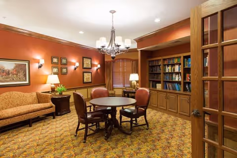 A cozy senior living common area with a round wooden table surrounded by four leather chairs. The room features a patterned carpet, a wooden bookshelf filled with books, a beige sofa, framed artwork on the walls, and warm lighting from wall sconces and table lamps. A chandelier hangs from the ceiling, and wooden blinds cover the window.