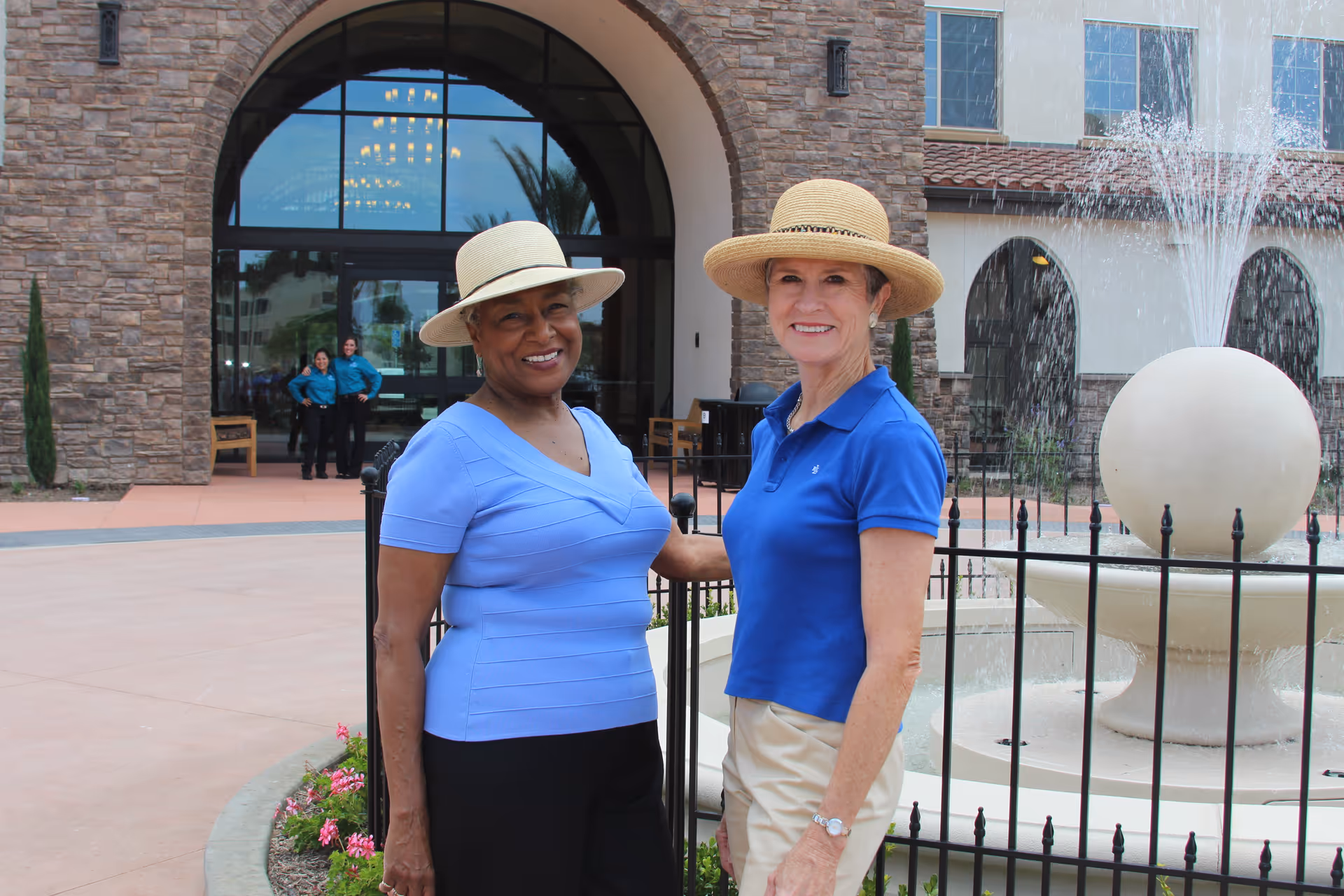 Two elderly women wearing hats and smiling, standing outside near a fountain with water spraying. Behind them is the entrance to a building with stone walls and large glass doors, with two staff members in blue uniforms standing in the background.