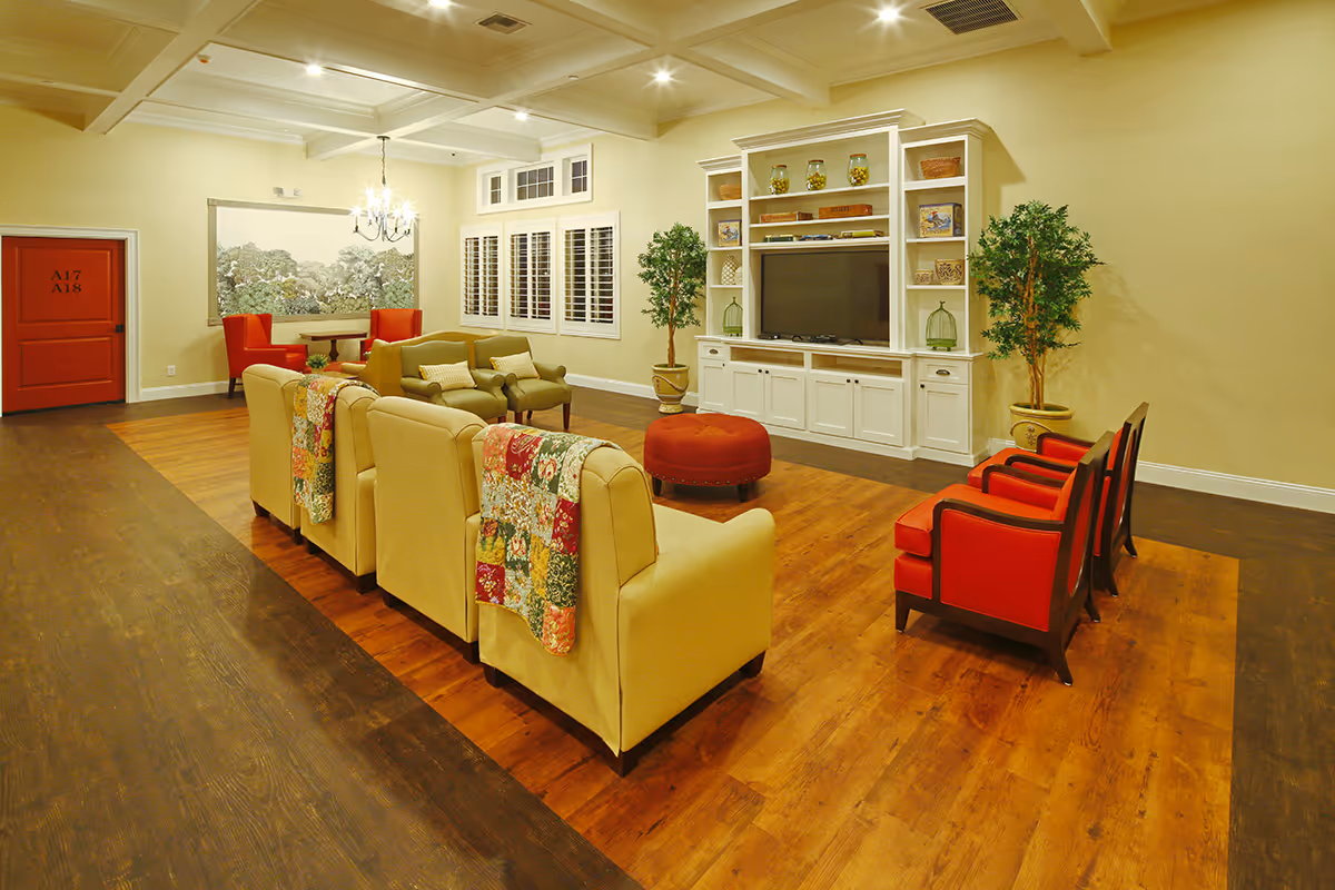 A spacious living room in an assisted living facility featuring a row of beige armchairs with colorful quilts draped over the backs, three red armchairs, a large white entertainment center with a flat-screen TV, decorative shelves, two potted plants, a round red ottoman, and a small table with two red chairs near a red door labeled A17 A18. The room has wooden flooring, cream-colored walls, and a chandelier hanging from the ceiling.