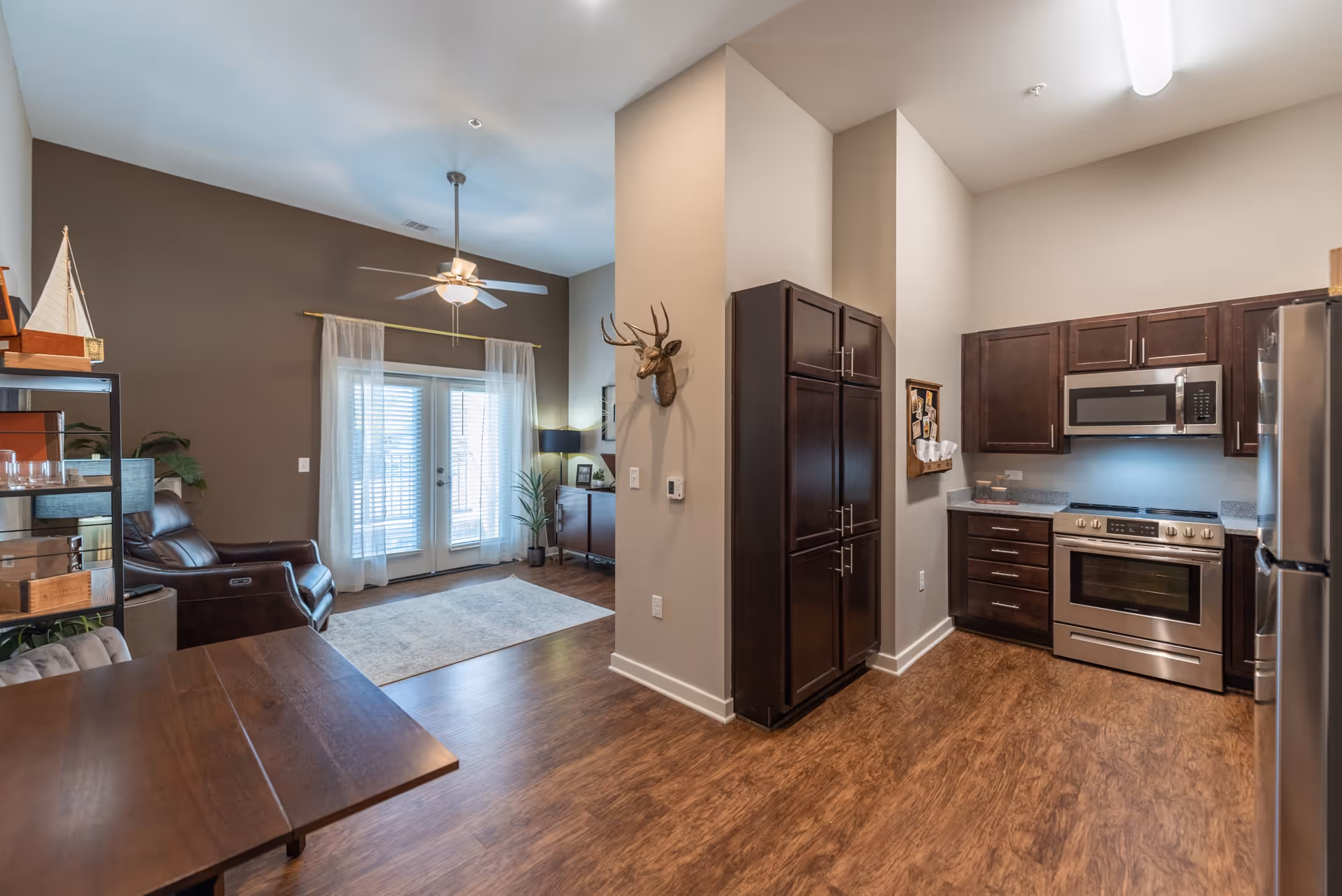 Interior view of a senior living facility apartment showing a kitchen area with dark wood cabinets, stainless steel appliances including a stove and microwave, and a refrigerator. Adjacent to the kitchen is a living area with a brown leather armchair, a ceiling fan, sheer curtains on glass doors, a rug, and decorative items including a mounted deer head on the wall.