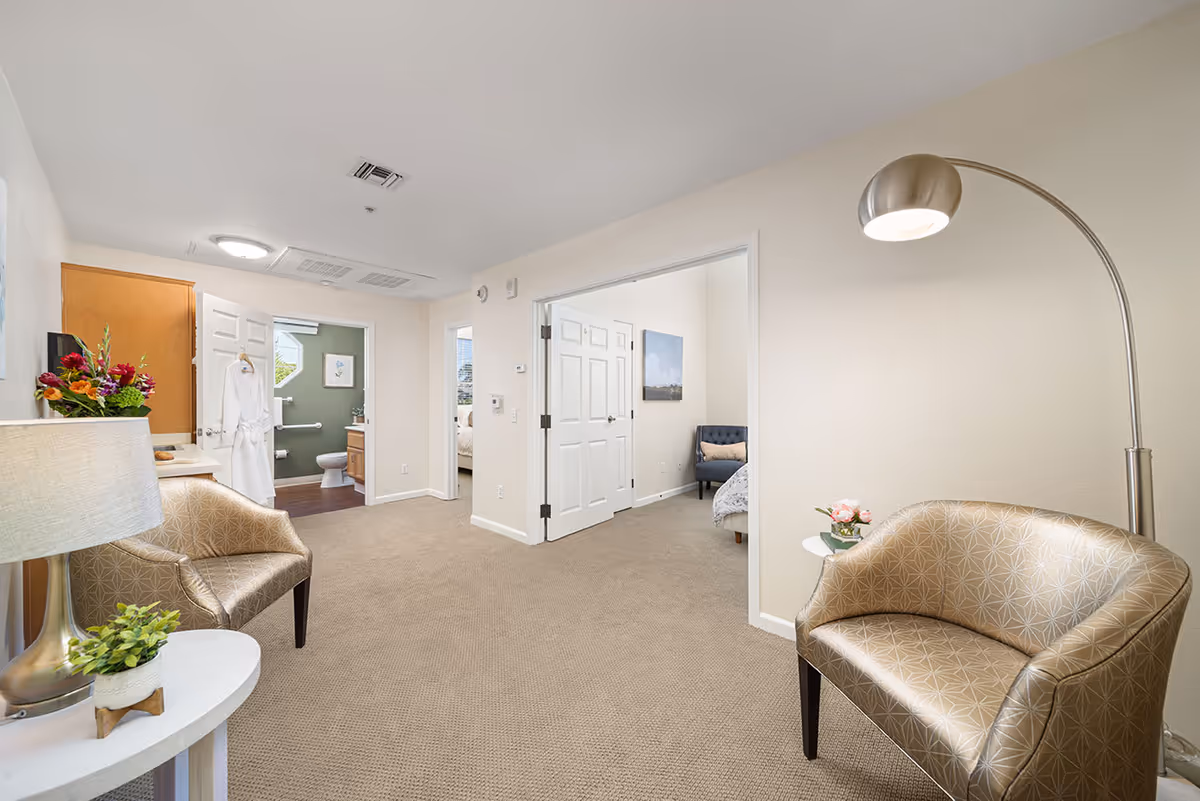 Interior view of a senior living facility room with beige carpet and light-colored walls. The room features two patterned armchairs, a floor lamp, a small round table with a plant, and an open doorway leading to a bedroom and a bathroom. The bathroom has a white robe hanging on the door and visible toilet and sink fixtures.