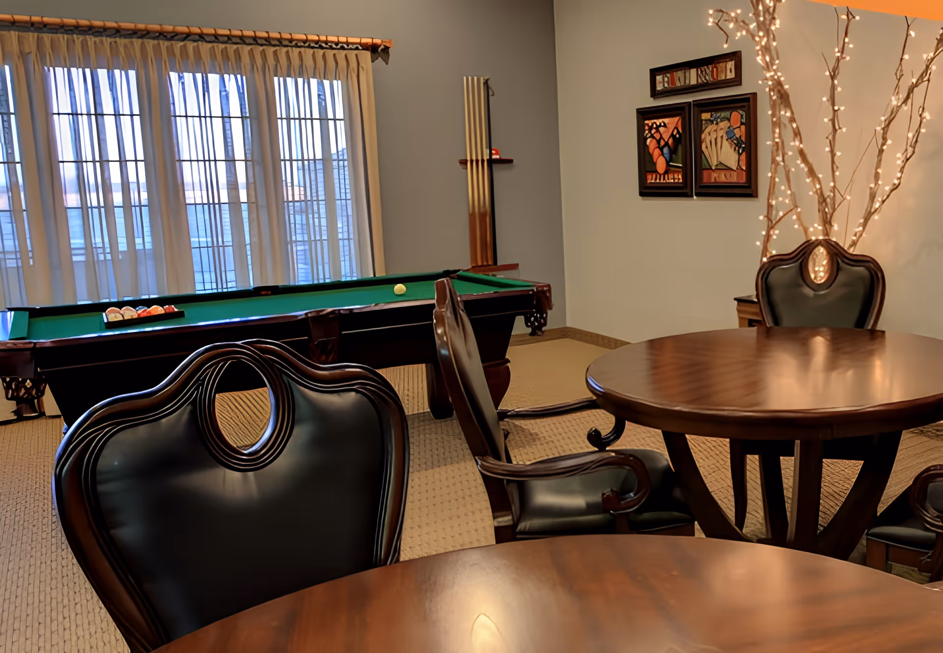 Interior room with a pool table near a window with curtains, two round wooden tables with dark leather chairs, and a decorative tree with string lights in the corner. There are framed pictures on the wall.