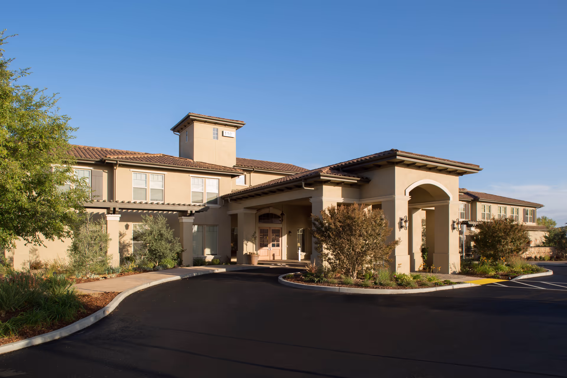 Exterior view of a senior living facility building with beige stucco walls, a tiled roof, and a covered entrance. The driveway curves in front of the entrance, and there are landscaped bushes and trees around the building under a clear blue sky.