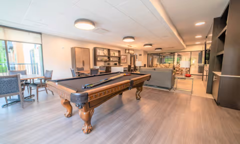 Well-lit communal lounge featuring a central pool table, sofas, and dining-style tables near large windows.