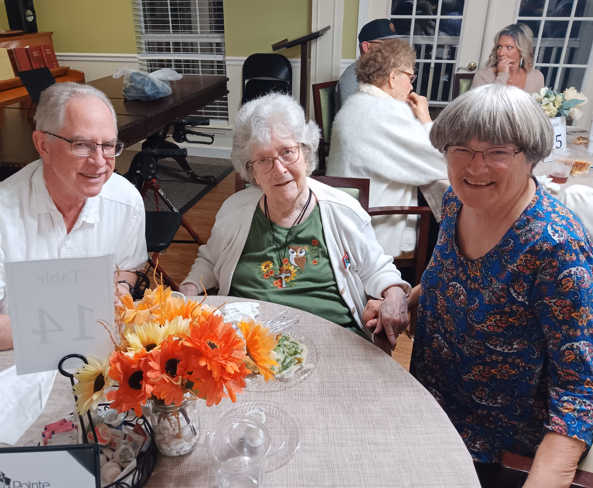 Three smiling older adults seated around a dining table with a bright floral centerpiece and a table number in a senior living dining room.