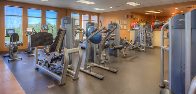 A well-lit fitness room with various exercise machines including stationary bikes, weight machines, and stability balls. Large windows on the left side allow natural light to fill the room, and framed pictures are visible on the far wall.