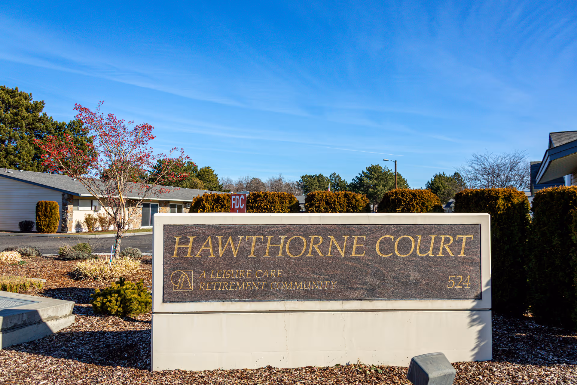 Outdoor view of the entrance sign for Hawthorne Court, a leisure care retirement community, with buildings, trees, and a clear blue sky in the background.