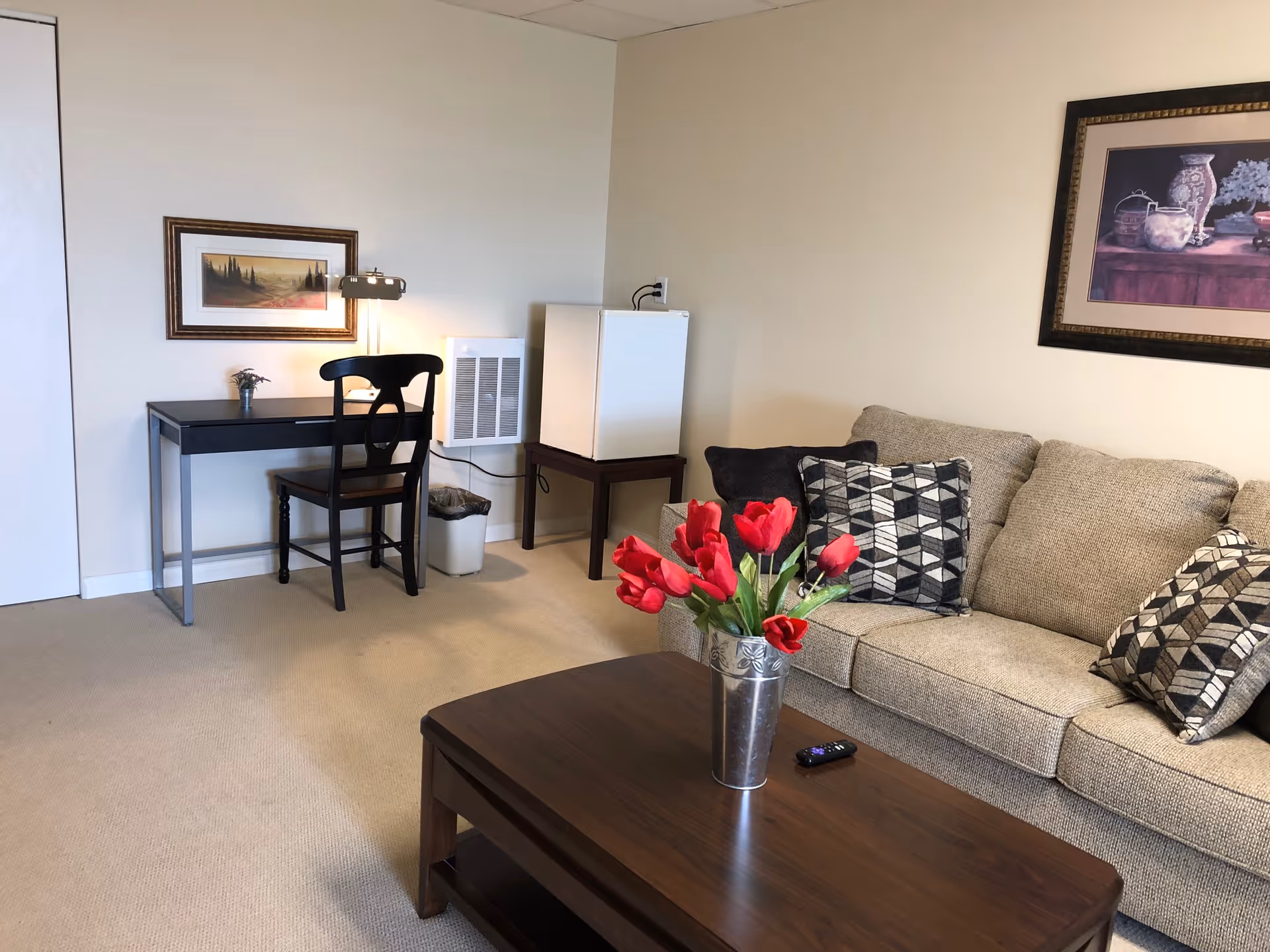 A cozy living room area with a beige couch adorned with patterned pillows, a wooden coffee table with a vase of red tulips and a remote control on it. To the left, there is a black desk with a matching chair, a small lamp, and a framed landscape painting on the wall. A small white refrigerator sits on a wooden stand next to the desk, and a trash bin is placed underneath the desk.
