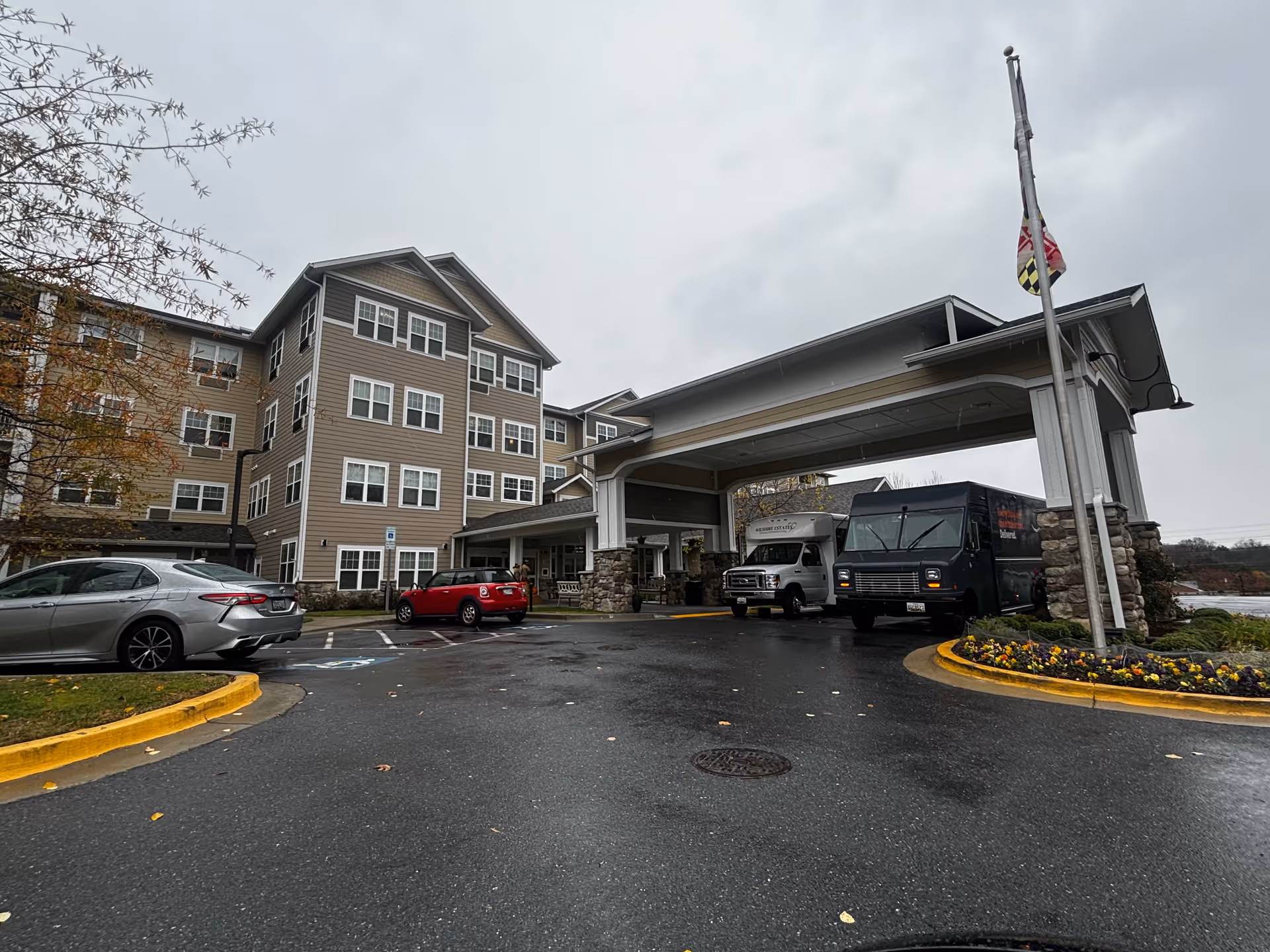 Exterior view of Wilshire Estates Gracious Retirement Living building on a cloudy day. The multi-story building has beige siding with white trim around numerous windows. There is a covered entrance with stone pillars and a flagpole flying a flag. Several vehicles, including a silver car, a red car, and two delivery trucks, are parked near the entrance. The ground is wet, indicating recent rain, and there are flower beds with yellow and purple flowers near the entrance.