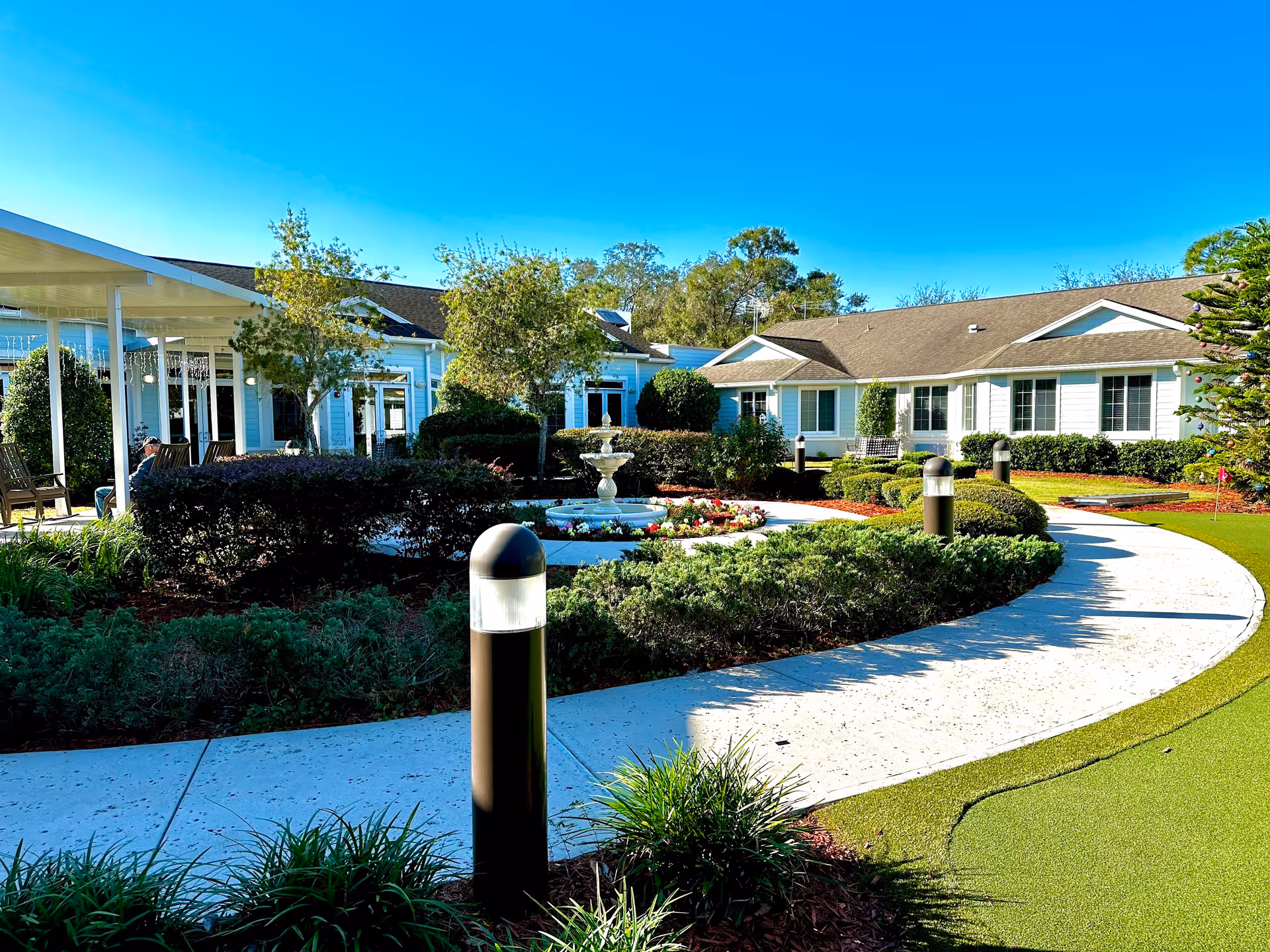 Curved paved path through a landscaped courtyard with a central fountain and one-story senior living buildings in the background.