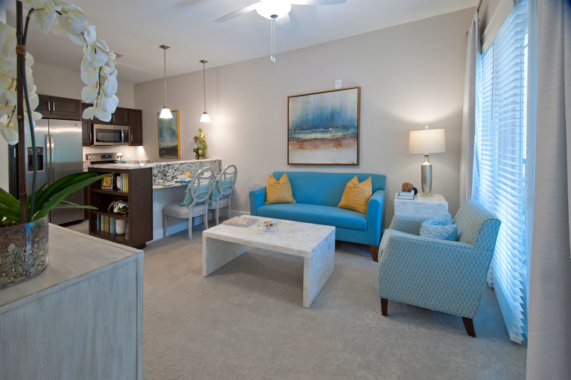 A cozy living room and kitchen area in a senior living facility. The living room features a blue sofa with two yellow pillows, a patterned armchair, a white coffee table, and a side table with a lamp. The kitchen area has dark wood cabinets, stainless steel appliances, a granite countertop with two bar stools, and pendant lights hanging above the counter. A large window with blinds allows natural light to fill the space, and there is a ceiling fan with a light fixture.