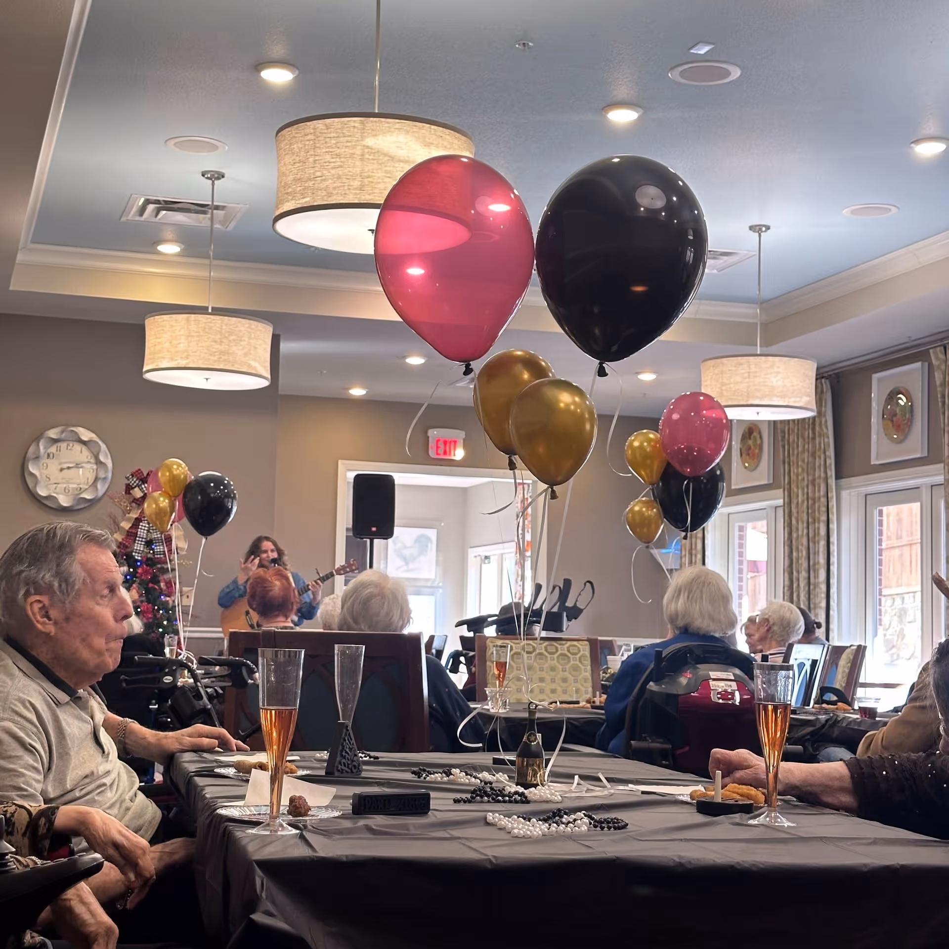 A group of elderly people seated around tables in a decorated common room with balloons in black, gold, and pink colors. A musician is playing a guitar in the background near a Christmas tree. The room has hanging ceiling lights and large windows with curtains.