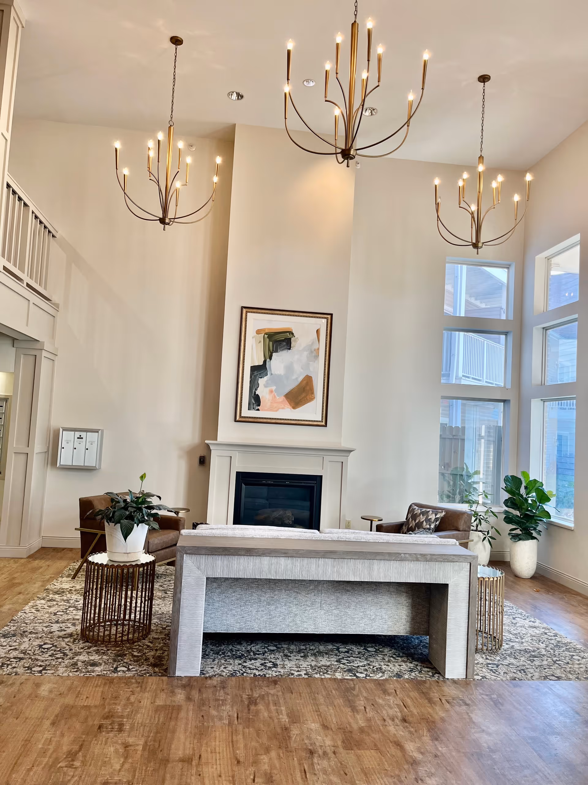 A bright and spacious living room area with high ceilings and three modern chandeliers. The room features a fireplace with abstract artwork above it, a light gray sofa facing the fireplace, two brown armchairs, side tables with potted plants, large windows letting in natural light, and a patterned area rug on wooden flooring.