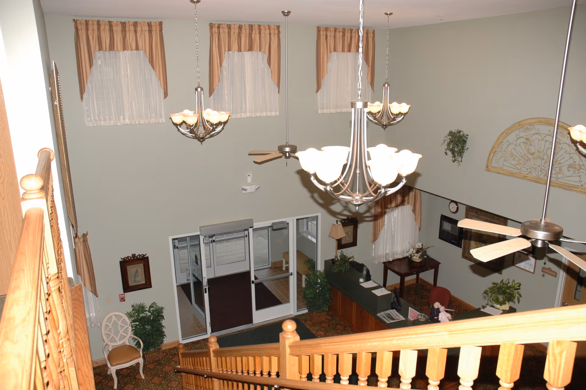 View from the top of a wooden staircase looking down into a lobby area with a reception desk, decorative plants, framed pictures on the walls, three chandeliers hanging from the ceiling, ceiling fans, and large windows with curtains. The entrance doors are glass and there is a chair and a small table near the walls.