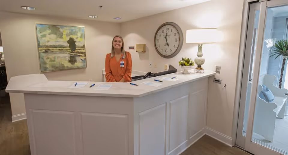 A smiling woman wearing an orange top stands behind a white reception desk in a well-lit room. The desk has papers and pens on it, and behind her is a large wall clock and a decorative lamp with a plant. A painting hangs on the wall to the left, and a glass door with a potted plant is visible on the right.