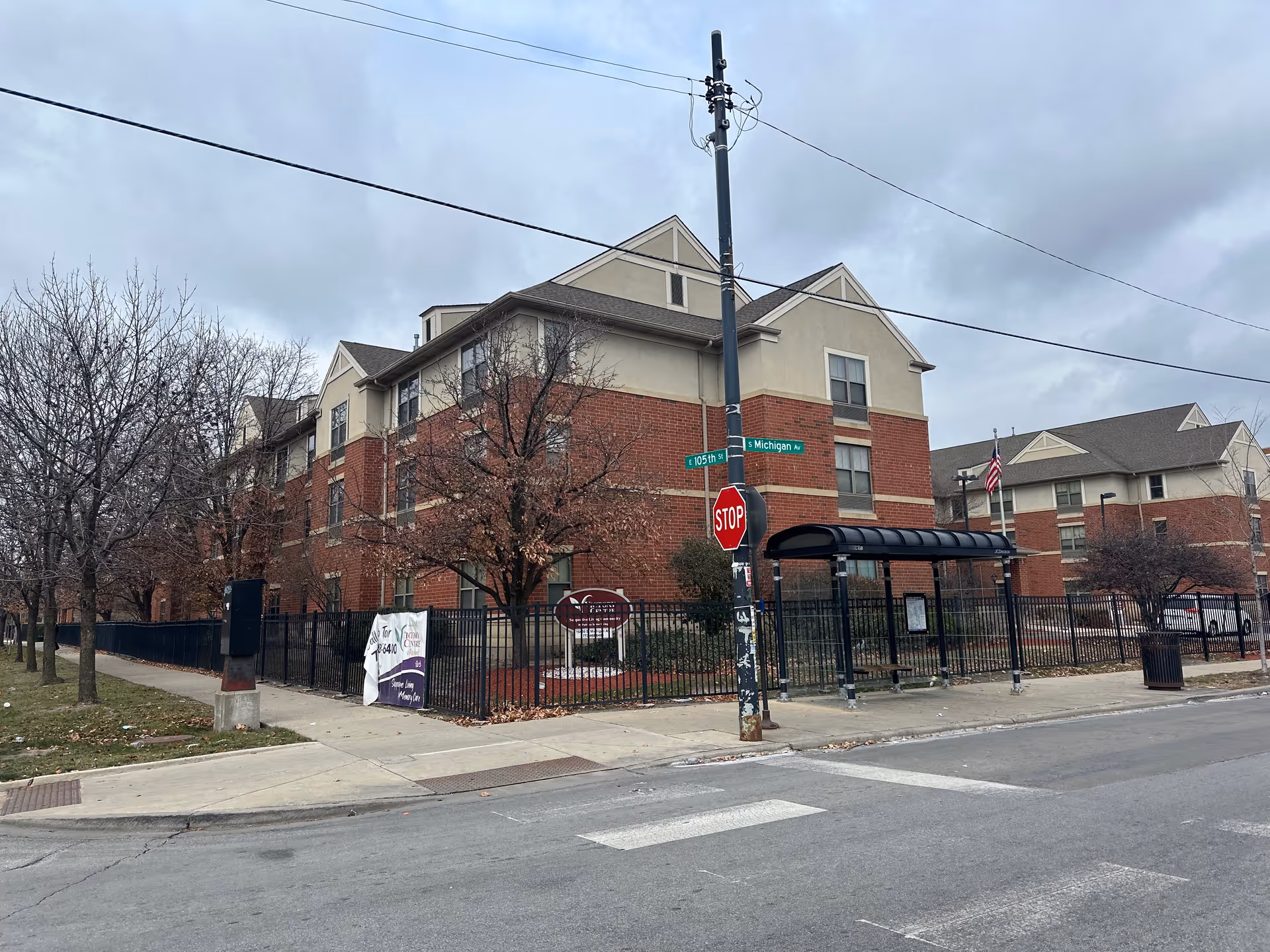 Three-story brick senior living building on a city corner with a fenced courtyard, stop sign, and covered bus shelter.
