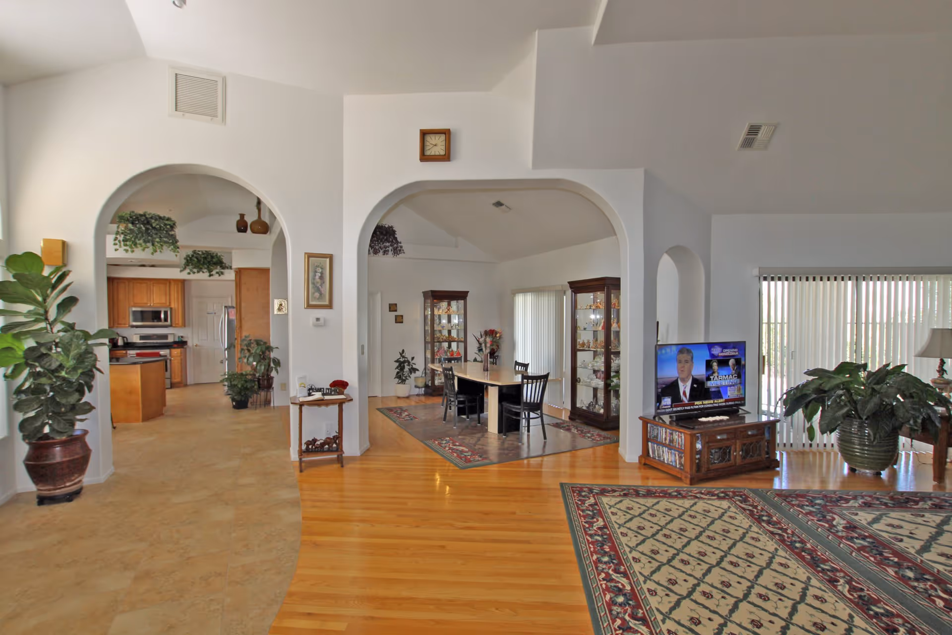 Interior view of a spacious assisted living facility showing a living room with a television on a wooden stand, a dining area with a table and chairs, and a kitchen with wooden cabinets. The floors are a mix of wood and tile, and there are several potted plants and decorative items throughout the space. Large windows with vertical blinds allow natural light to fill the room.