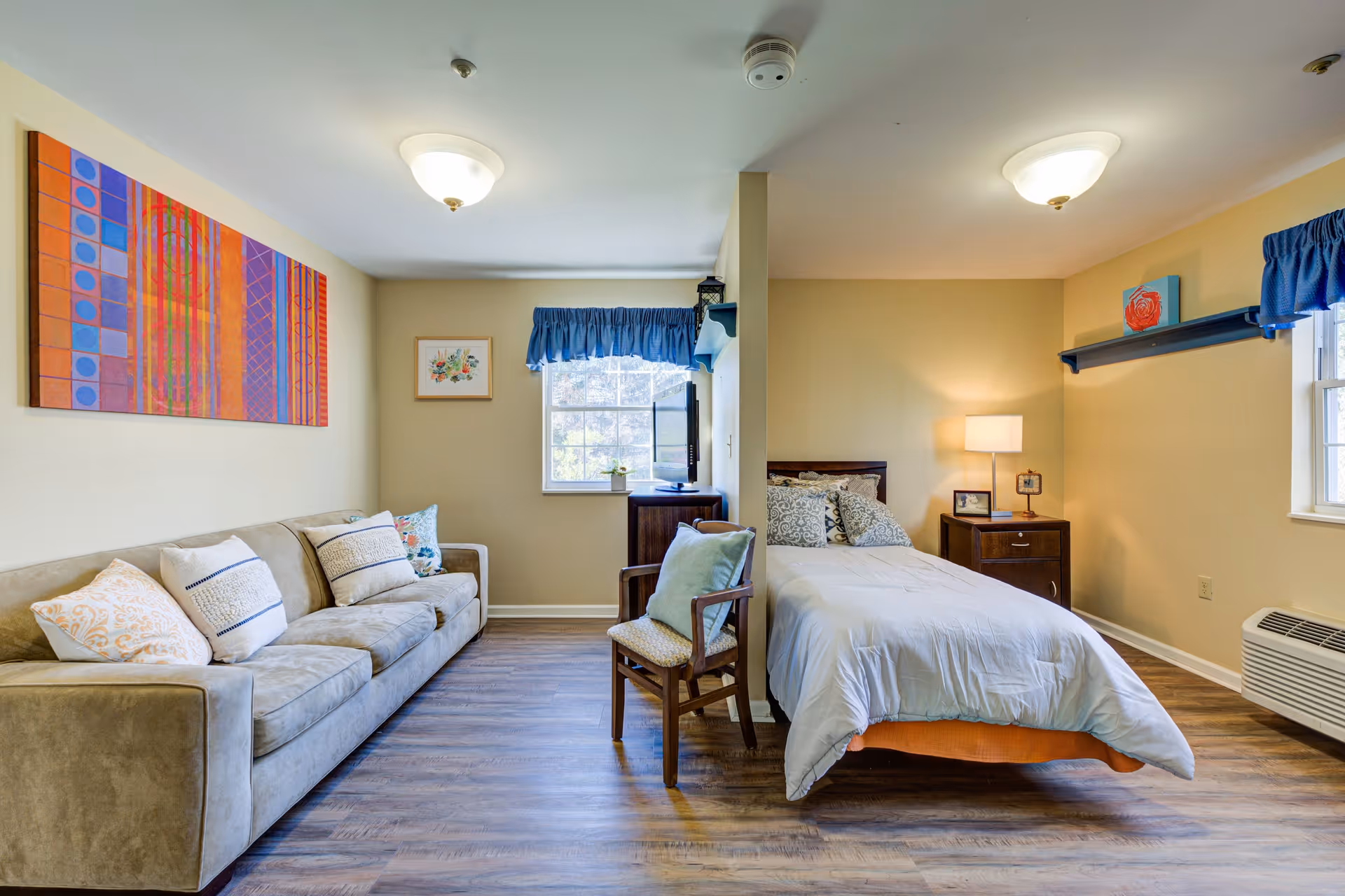 A cozy room divided by a partial wall, featuring a beige sofa with decorative pillows on the left side and a single bed with white bedding and patterned pillows on the right side. The room has wooden flooring, two windows with blue valances, a wooden chair with a cushion, a nightstand with a lamp and framed photos, and colorful artwork on the walls.