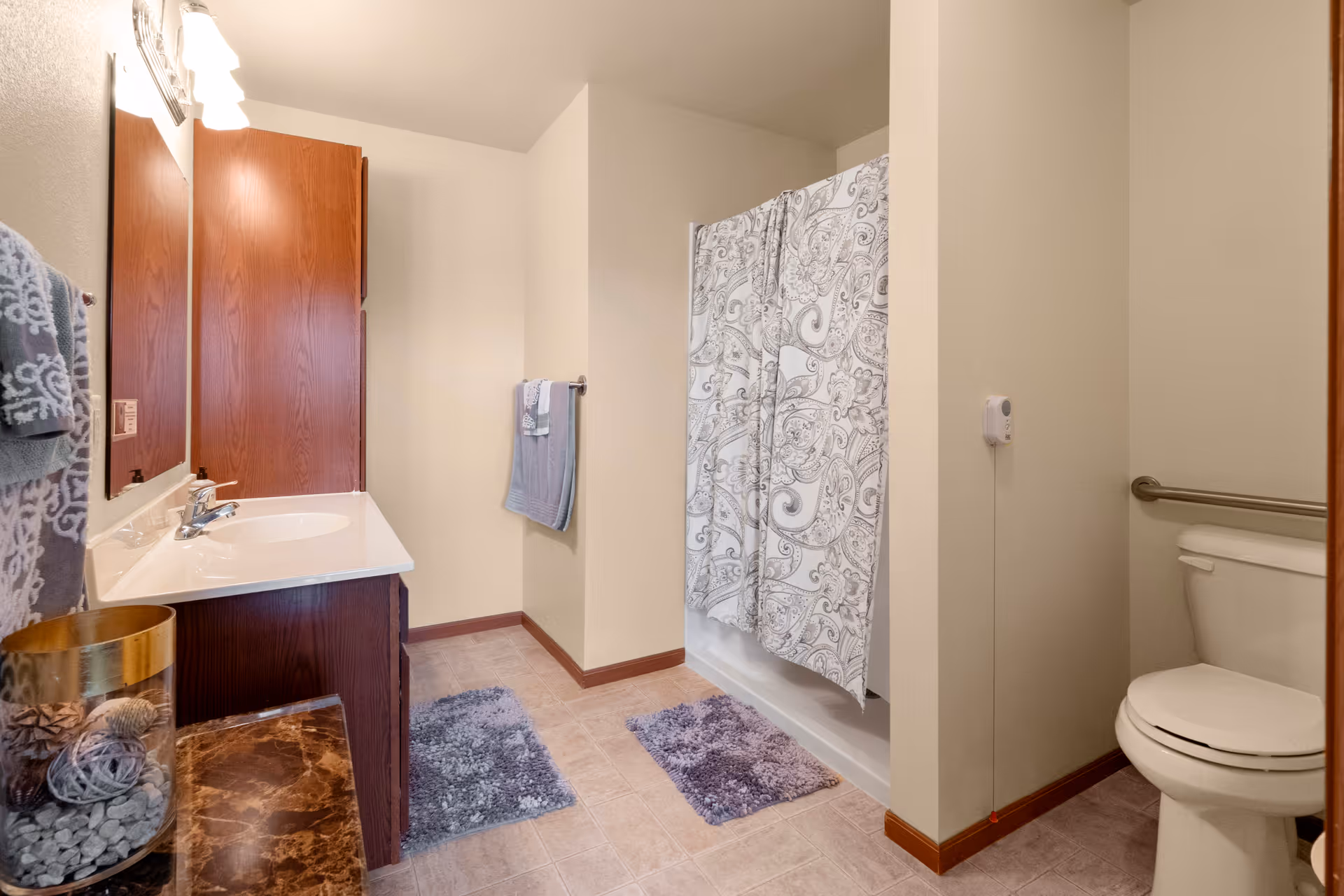 A clean and well-lit bathroom featuring a white sink with a wooden cabinet, a large mirror, a toilet with a grab bar, a shower with a patterned curtain, and two purple bath mats on the tiled floor. Towels hang on the wall near the sink and shower.