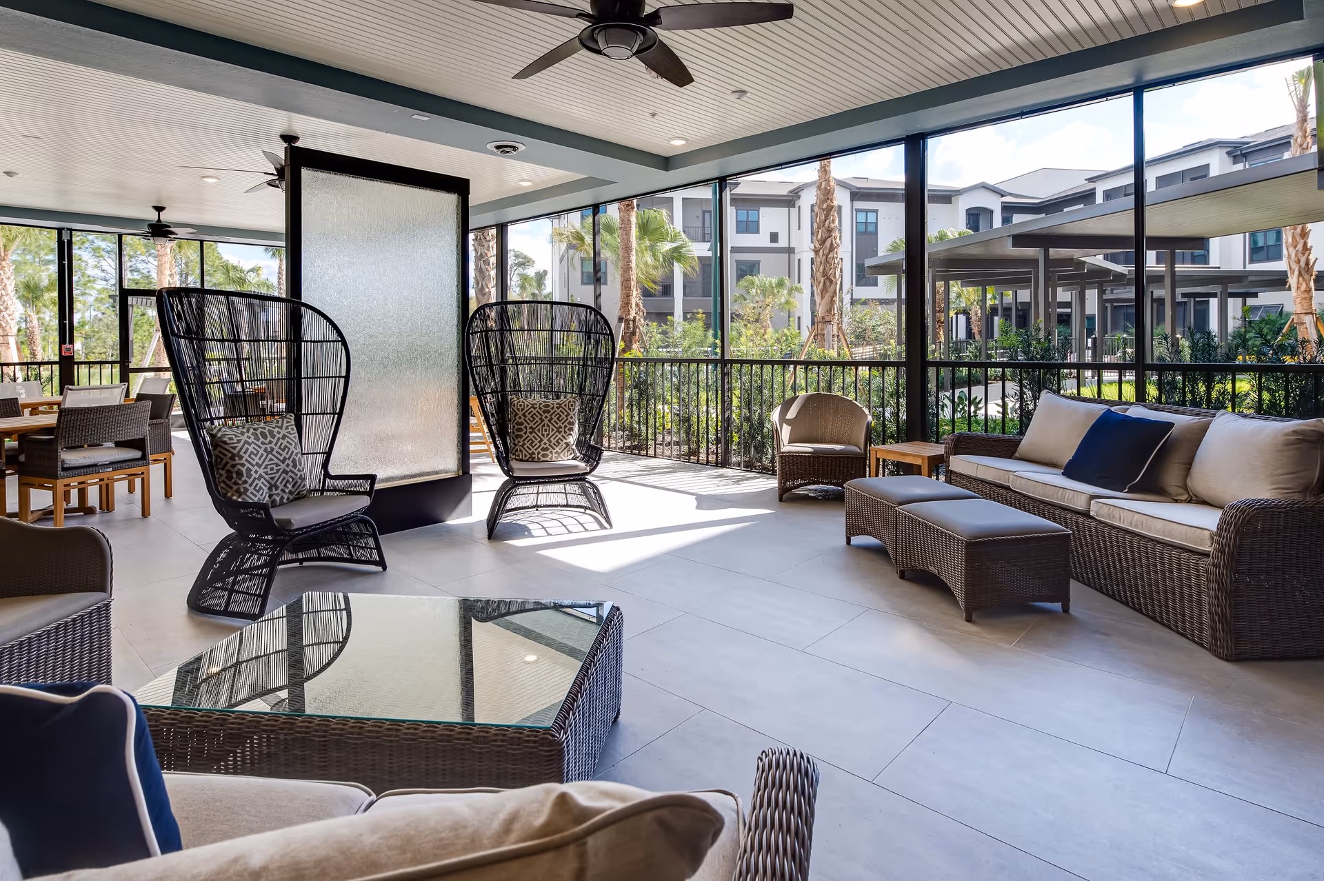 A spacious, covered outdoor lounge area with wicker furniture including cushioned sofas, armchairs, and a glass-top coffee table. The area is enclosed with large glass windows offering a view of palm trees and a modern building outside. Ceiling fans are mounted on the white paneled ceiling.