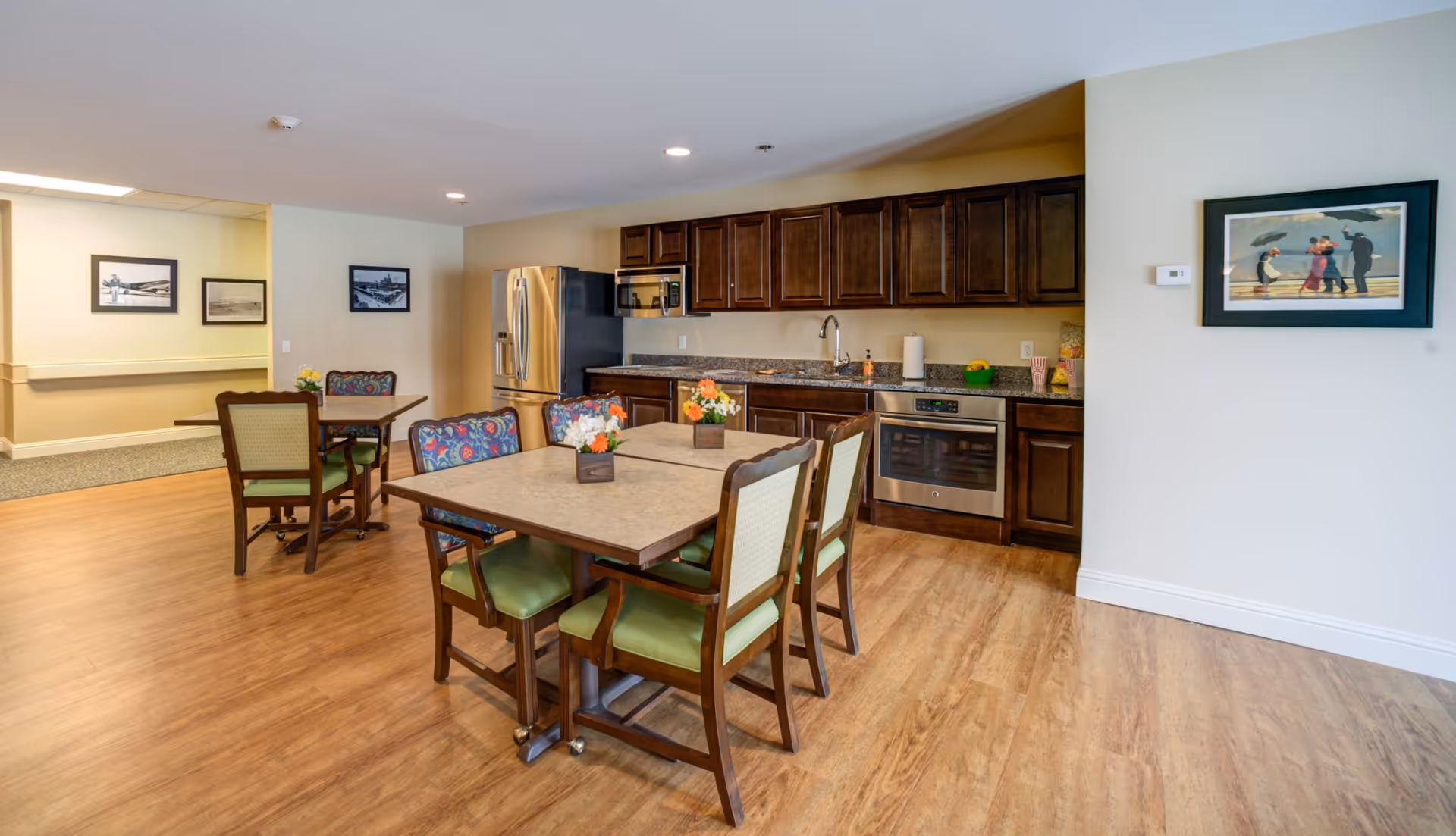A communal kitchen and dining area in Oak Pointe Assisted Living featuring wooden cabinets, stainless steel refrigerator, microwave, and oven. There are two tables with chairs around them, decorated with small flower arrangements. The room has wood flooring and framed artwork on the walls.
