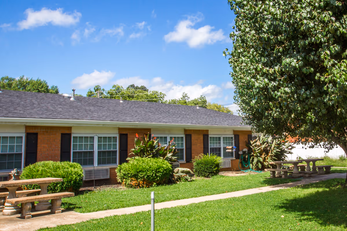 Exterior view of a single-story brick building with multiple windows, surrounded by green bushes and trees. There are concrete picnic tables and benches on a grassy area with a sidewalk running through it under a blue sky with some clouds.