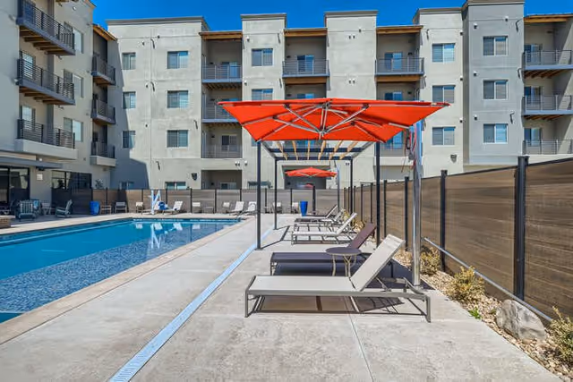 Outdoor pool area at 55 Resort Apartments Downtown Greeley with lounge chairs and red umbrellas alongside the pool, surrounded by a multi-story apartment building under a clear blue sky.