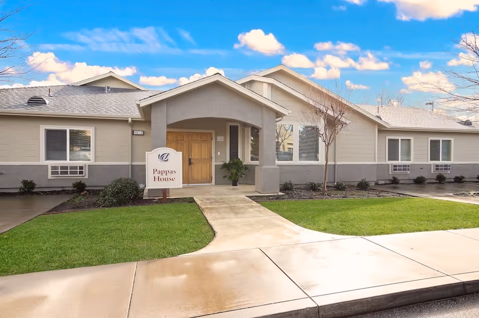 Exterior view of a single-story building with beige siding and a gray roof under a blue sky with clouds. A concrete walkway leads to a covered entrance with double wooden doors. A sign near the entrance reads 'Pappas House'. There is green grass and some small shrubs around the building.