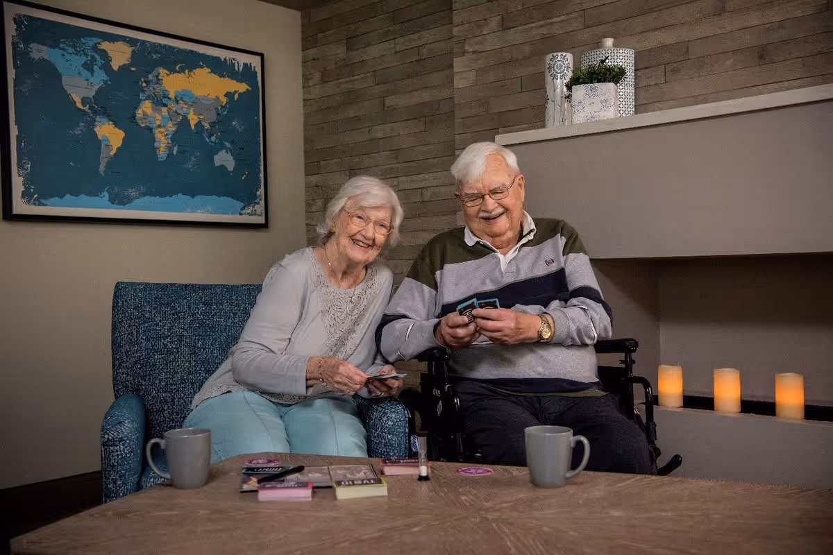 An elderly woman and man sitting together in a cozy living room area, smiling and playing cards. The woman is seated in a blue armchair, and the man is in a wheelchair. A world map is hanging on the wall behind them, and there are decorative candles and plants on a shelf next to a fireplace.