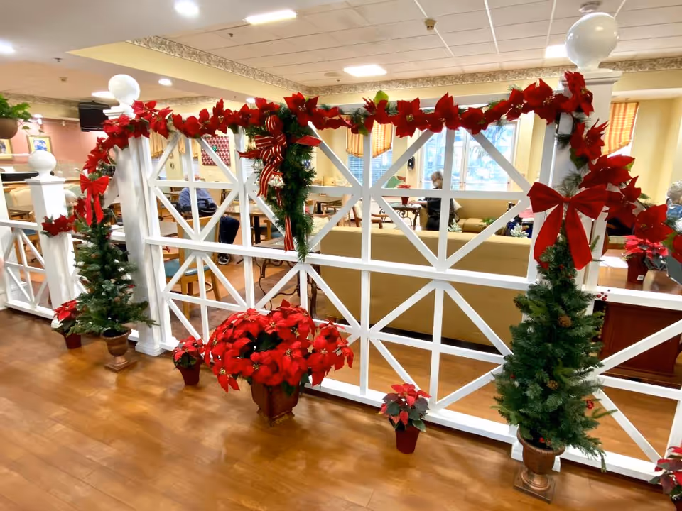 A decorated white partition in a senior living common area adorned with garlands, poinsettias, small Christmas trees and red bows with seating visible behind.