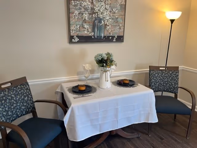 A small dining area with a table covered by a white tablecloth set for two. Each place setting has a dark plate with a muffin on it, a fork, and a knife. A white vase with flowers is centered on the table. Two chairs with wooden arms and patterned upholstery are positioned on either side of the table. A floor lamp is in the corner, and a floral painting hangs on the wall above the table.