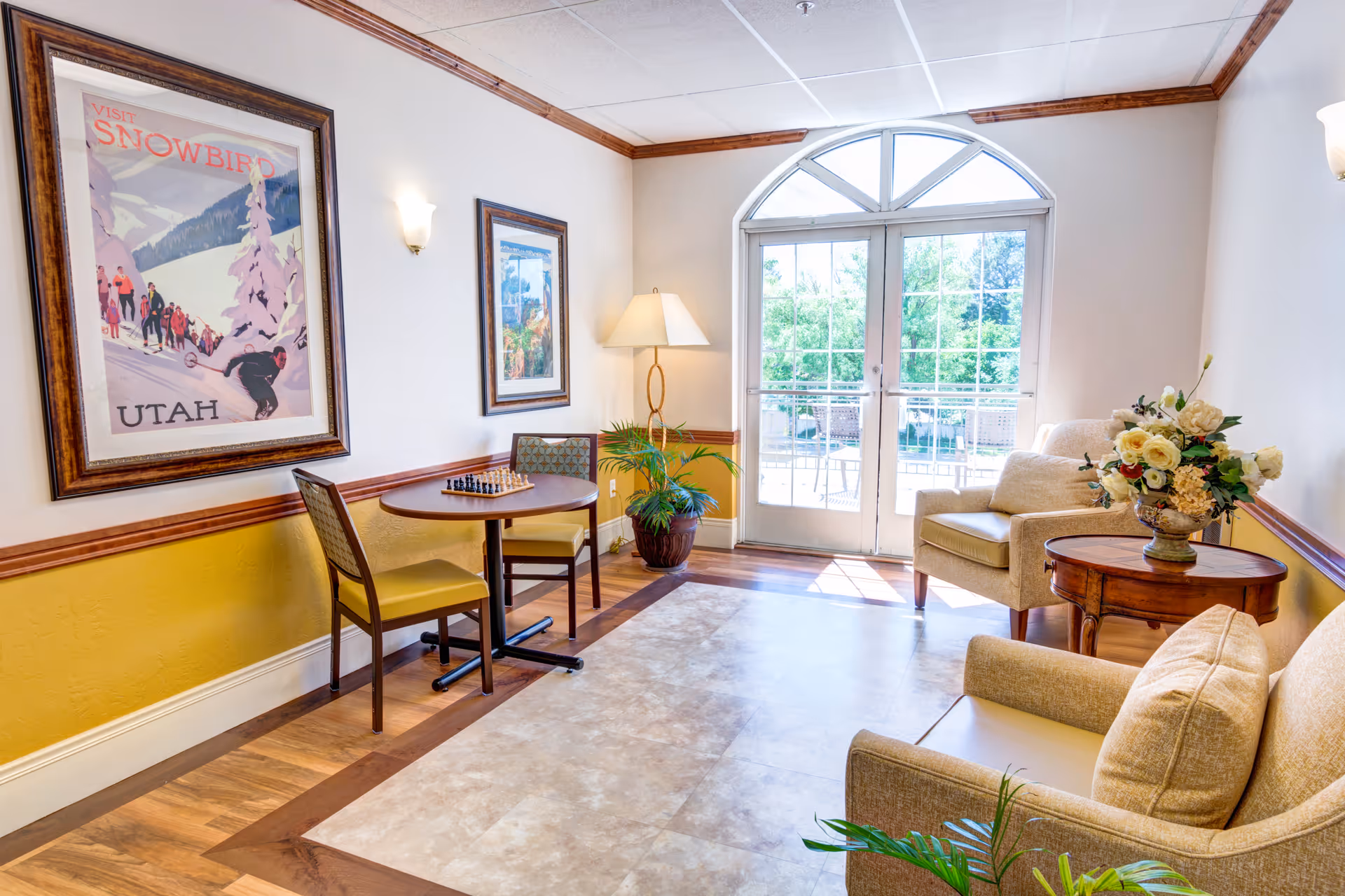 Bright seating area with armchairs, a small table with a chessboard, framed wall art, and French doors leading to a balcony.