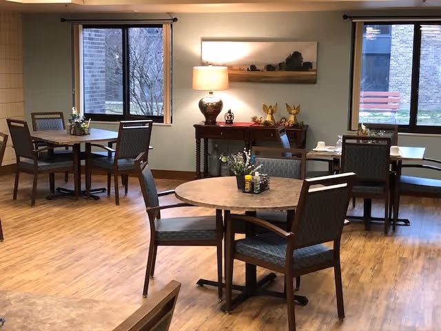 A dining area with several round and square tables, each surrounded by four chairs. The room has wooden flooring, two large windows showing an outdoor view, and a side table with a lamp, decorative items, and a landscape painting above it.
