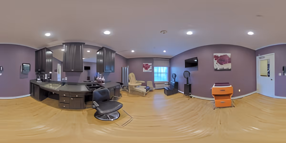 Panoramic view of a purple-walled interior common room with salon chairs, a counter with cabinets, seating and a wall-mounted TV.