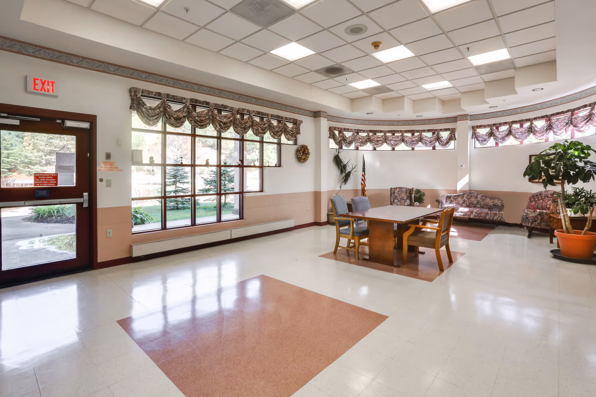 A bright and spacious common area in a senior living facility with large windows adorned with floral valances. The room features a table with four chairs in the center, floral patterned sofas along the curved wall, potted plants, and an American flag. There is an exit door with a red exit sign above it on the left side.