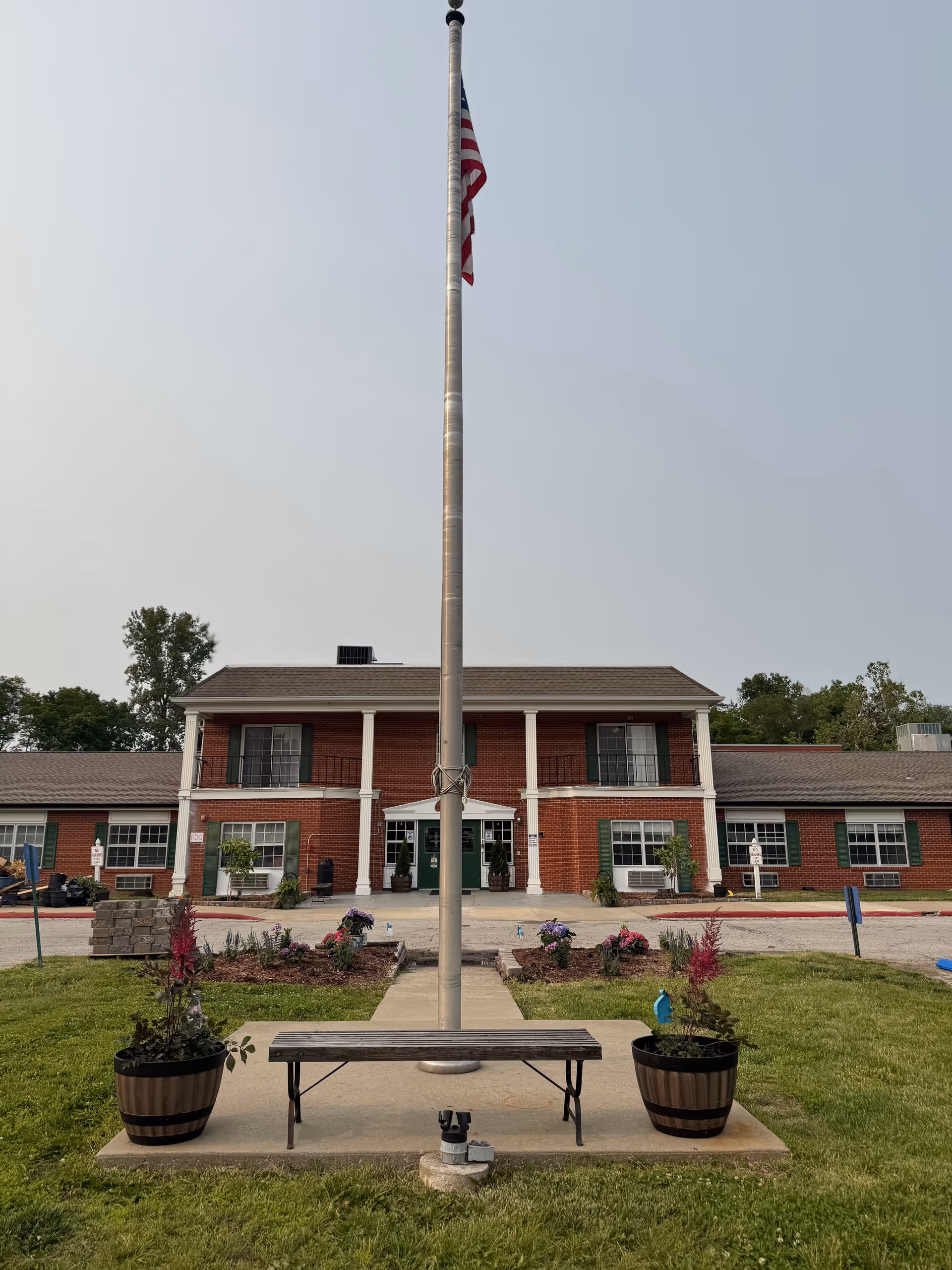 Front view of a two-story brick building with an American flag on a tall flagpole and a bench with planters in the foreground.