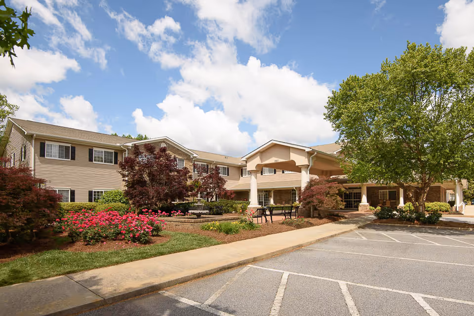 Front exterior of a senior living building with a covered entrance, landscaped gardens, and a parking area under a blue sky.