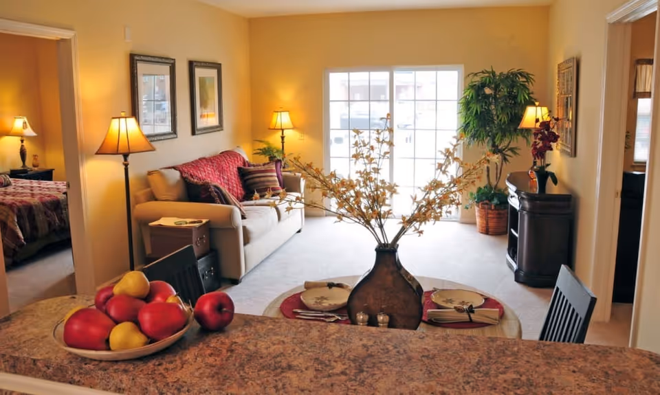 A cozy living room with a beige sofa adorned with red and striped pillows, two framed pictures on the wall, and two floor lamps providing warm lighting. A round coffee table in front of the sofa holds a decorative vase with branches and two place settings. To the left, a bedroom with a bed covered in a red and brown quilt is visible through an open doorway. A large window or sliding glass door at the back lets in natural light. In the foreground, a countertop with a bowl of red and yellow apples is visible. There are also plants and a dark wooden cabinet on the right side of the room.
