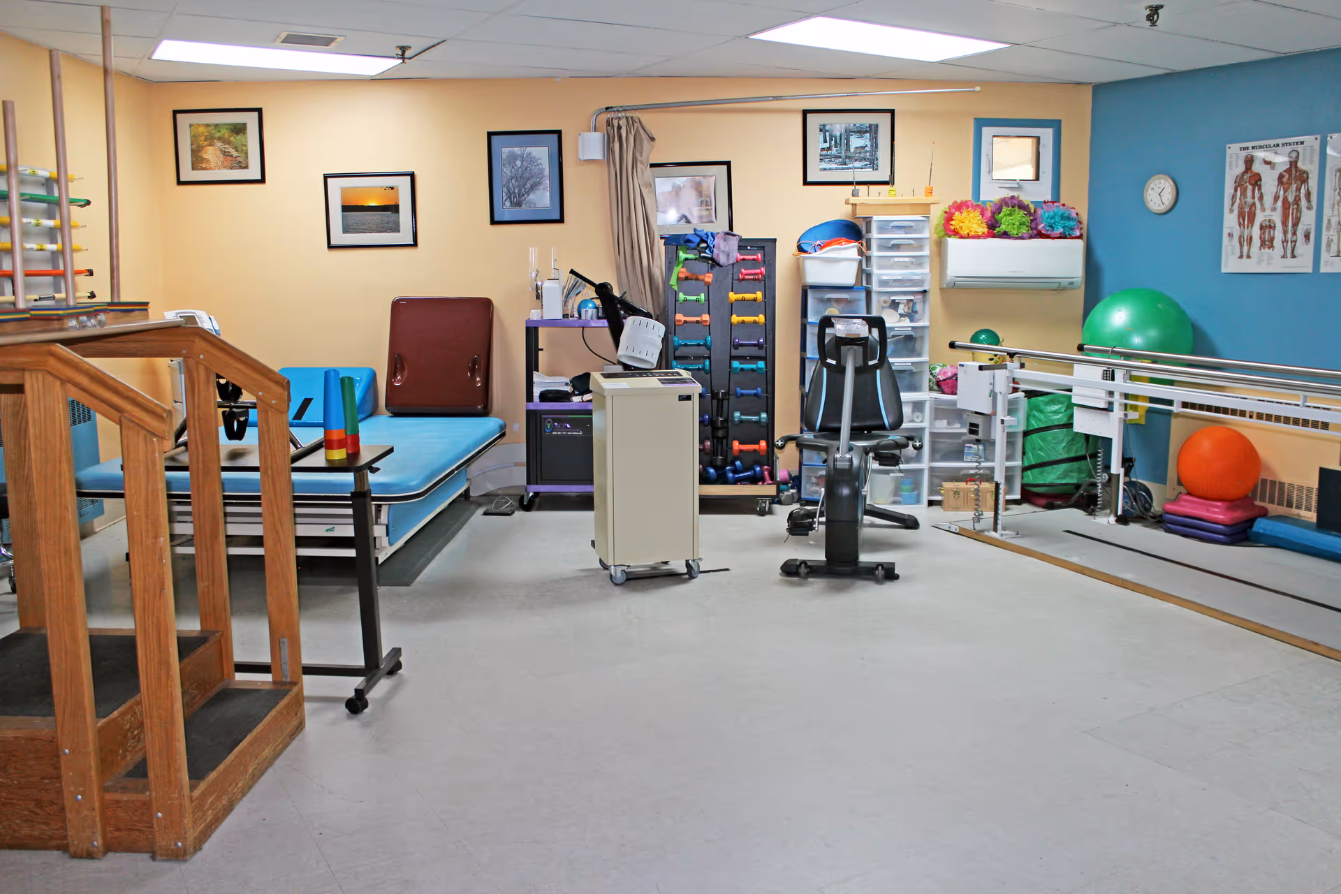 A physical therapy room in a care facility featuring wooden stairs with handrails, a padded therapy table, exercise equipment including a stationary bike, dumbbells, therapy balls, and parallel bars. The walls are decorated with framed pictures and anatomical charts.
