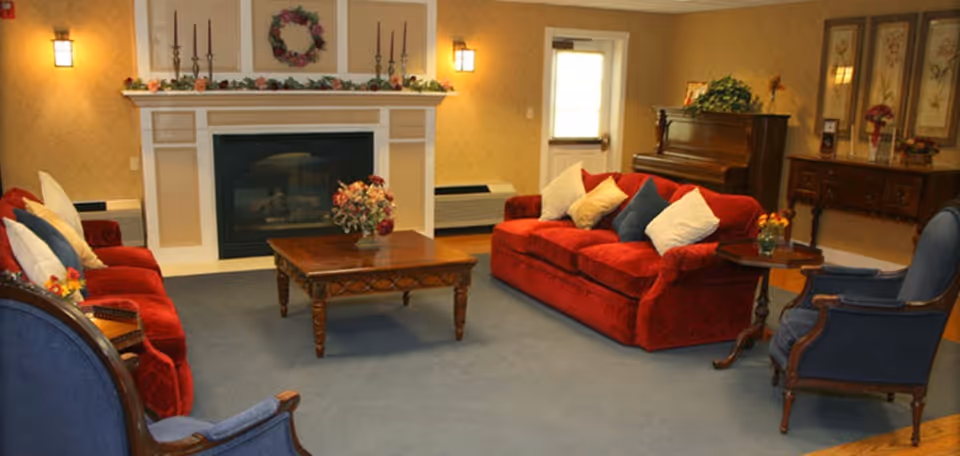 A cozy living room in an assisted senior living facility featuring two red sofas with multiple cushions, two blue armchairs, a wooden coffee table with a floral centerpiece, a fireplace decorated with a wreath and candles, a piano, and a wooden sideboard with framed artwork and flowers.