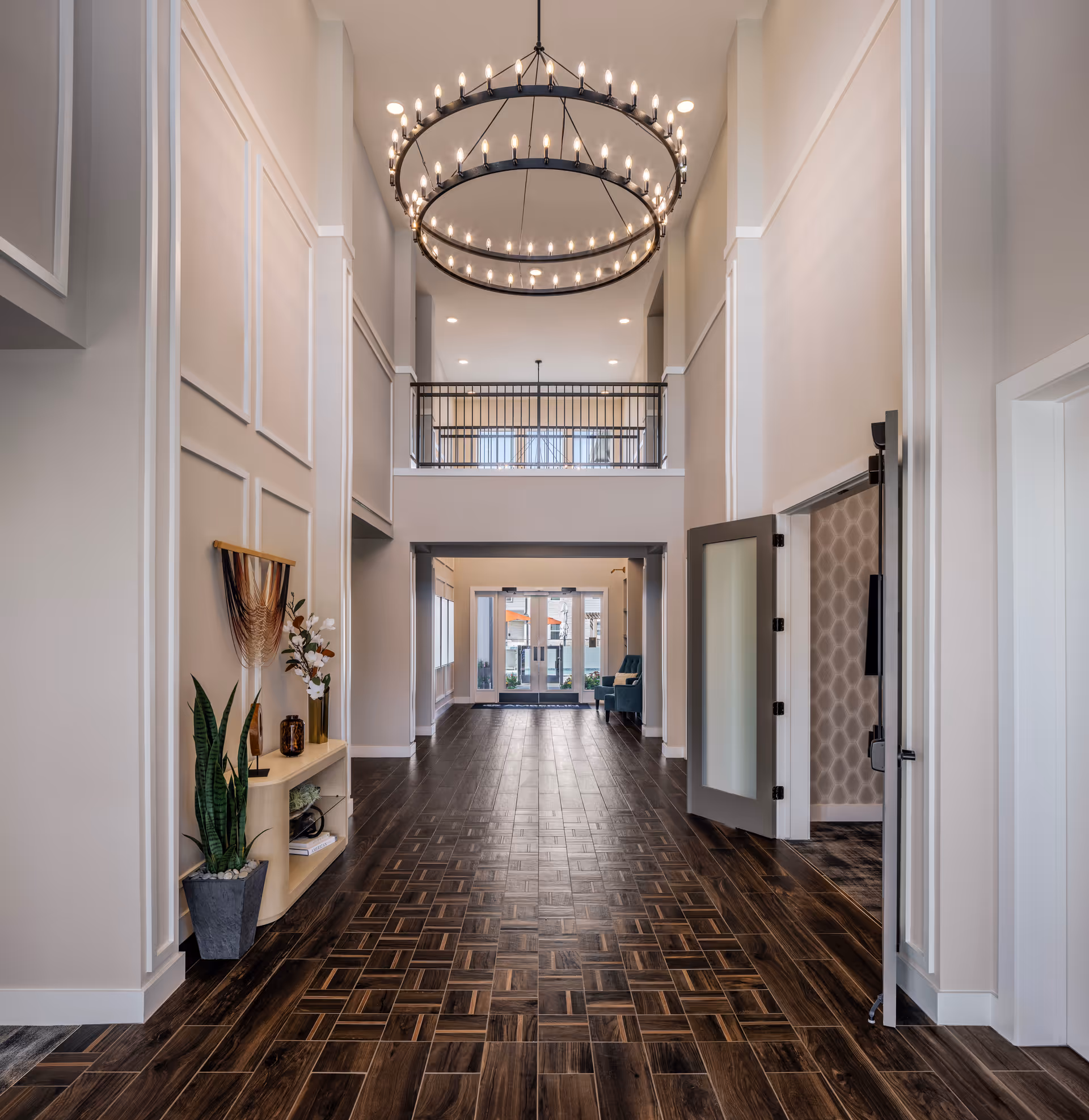 Bright two-story lobby interior with a large circular chandelier, patterned wood-look tile floor, decorative shelving and seating area at the far end.