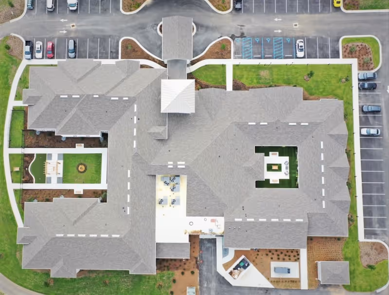 Aerial view of The Canopy at Walden Woods senior living facility showing the building's roof, parking lots with cars, landscaped green areas, and outdoor seating spaces within the building's courtyard.