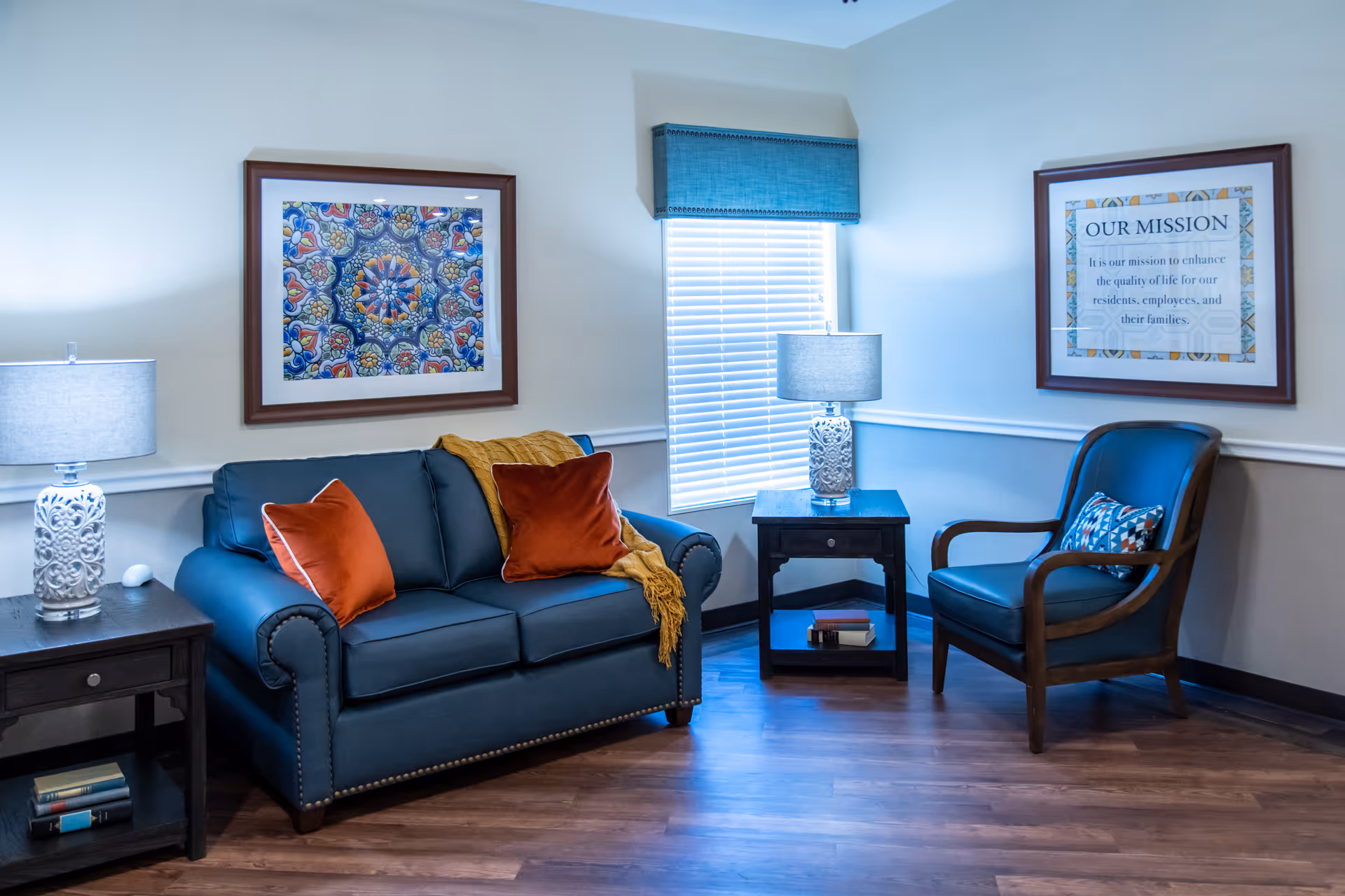 A cozy sitting area in a nursing and rehabilitation center featuring a blue loveseat with two orange pillows and a yellow throw blanket, a wooden armchair with a patterned cushion, two side tables each with a decorative lamp, framed artwork on the walls, and a window with blinds and a blue valance.