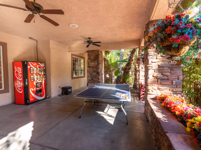 Covered outdoor patio area with a ping pong table in the center, a Coca-Cola vending machine against the wall, ceiling fans, and colorful hanging and planter flowers along a stone wall with palm trees visible outside.