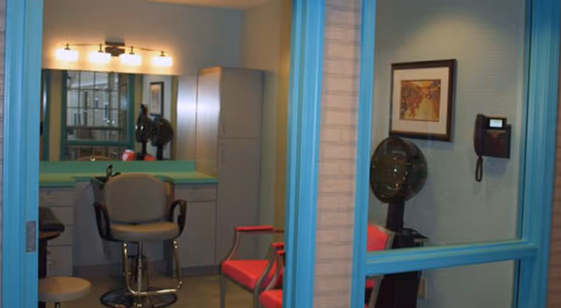 Small salon/styling room seen through turquoise-framed interior windows with a salon chair at a mirror, a hooded hair dryer, and red waiting chairs.