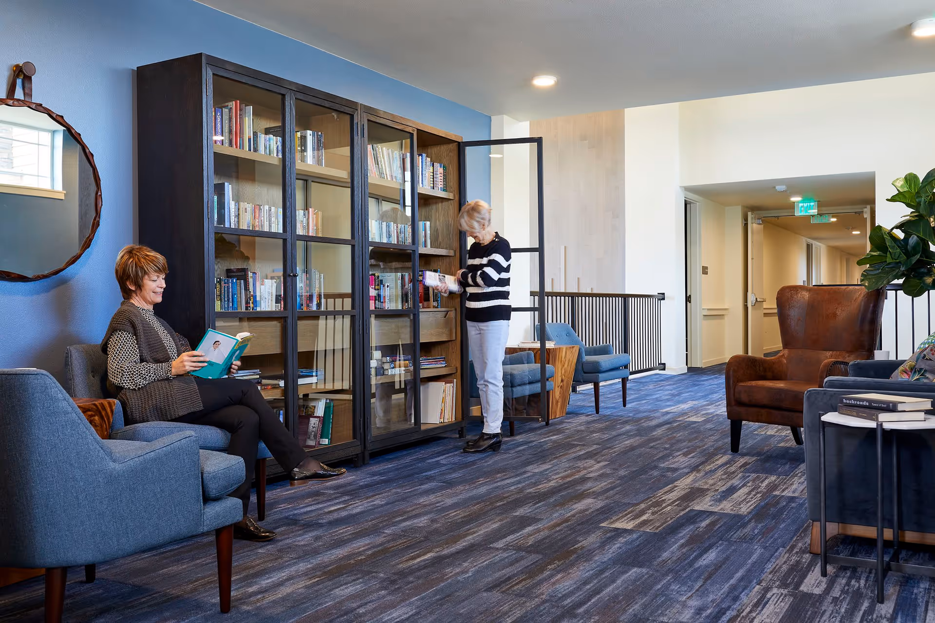 A cozy reading area in a senior living facility with two women. One woman is sitting comfortably in a blue armchair reading a book, while the other woman is standing and selecting a book from a tall glass-front bookshelf. The room features blue walls, a round mirror, blue patterned carpet, additional seating including a brown leather armchair, and a small side table with books.